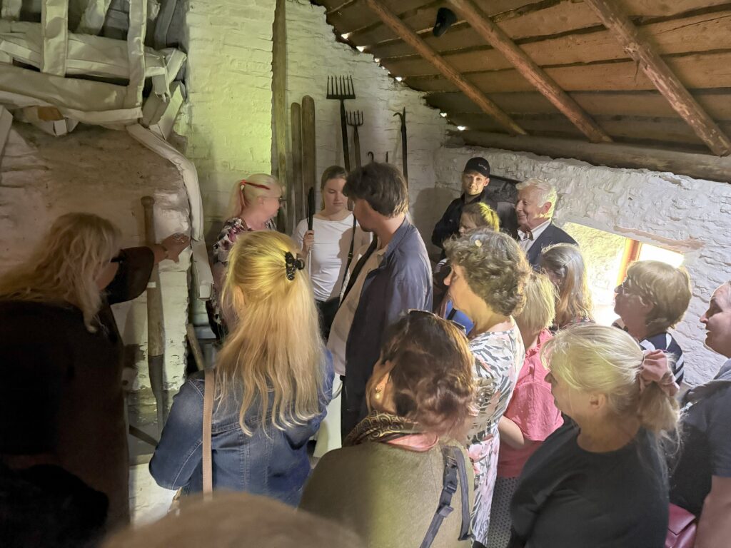 nüüd on grupp jõudnud tuppa, puidust seintega taluruumi, mille seintel erinevad tööriistad, rehadest vikatiteni. Kuulavad giidi.