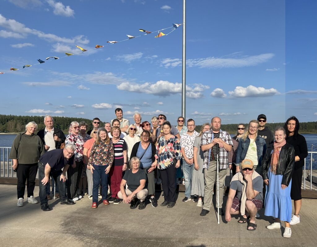 Jälle grupipilt, 20 inimest seismas värviliste lippude taustal, kivisillutisel.