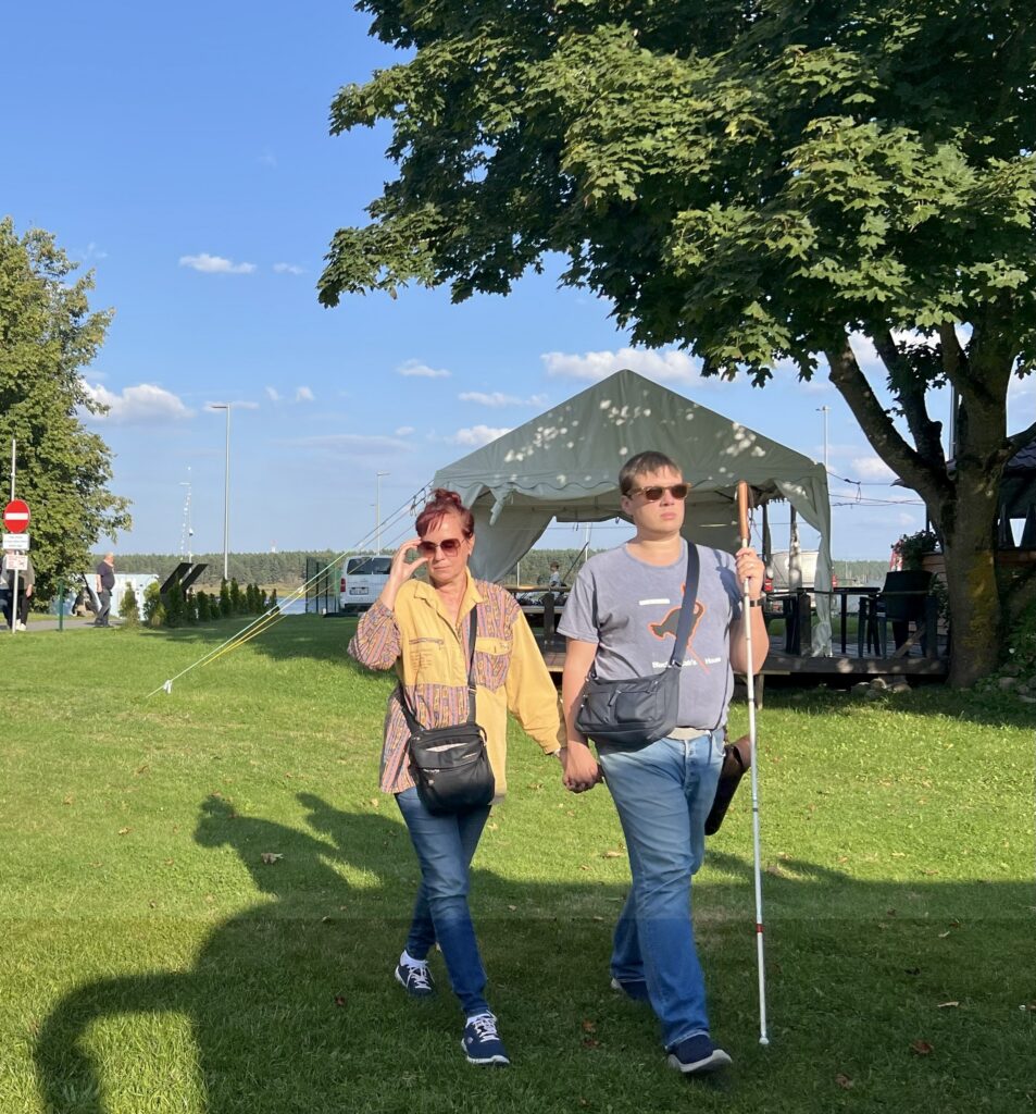 Kaks inimest, mees ja naine käsikäes looduses jalutamas. Mehel on käes valge kepp ja peas päikeseprillid.