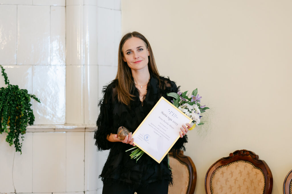 Anna-Aurelia seisab valgusküllases ruumis, käes lillekimp ja tänukiri. Tal on sirged pruunid juuksed ning ta kannab musta mustrilist pluusi ja pükse.