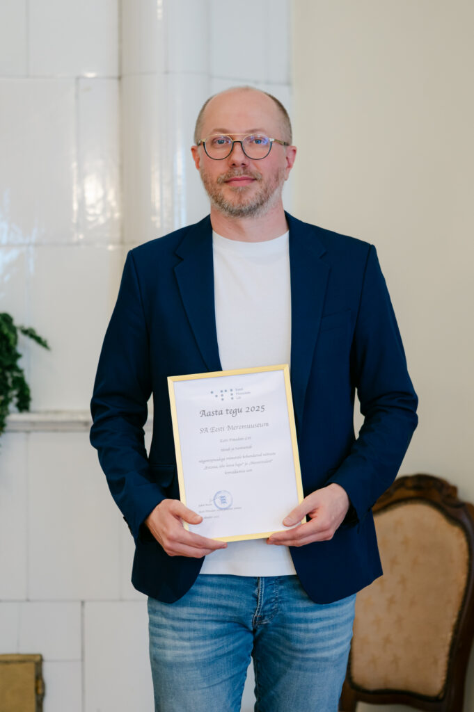 Mihkel seisab heleda seina ees, käes Aasta Tegu 2025 tänukiri. Tal on tumesinine pintsak, valge särk ja heledad teksad.