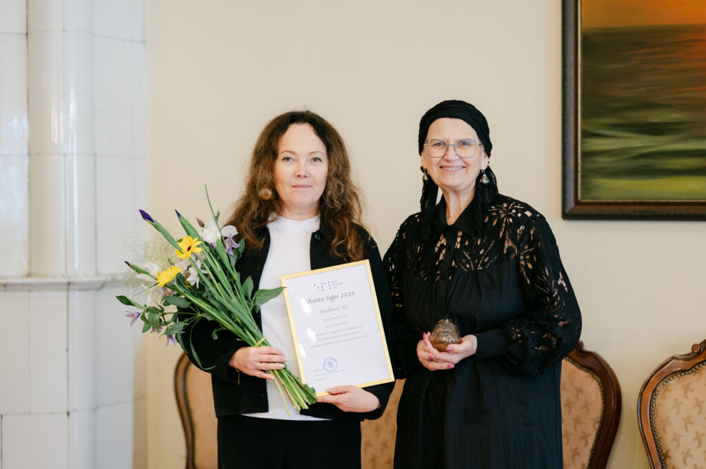 Kaks naist seisavad kõrvuti, üks hoiab käes lillekimpu ja tänukirja, teine väikest klaasist aasta teo meenet. Mõlemad on riietatud musta ning naeratavad kaamerasse sõbralikult.